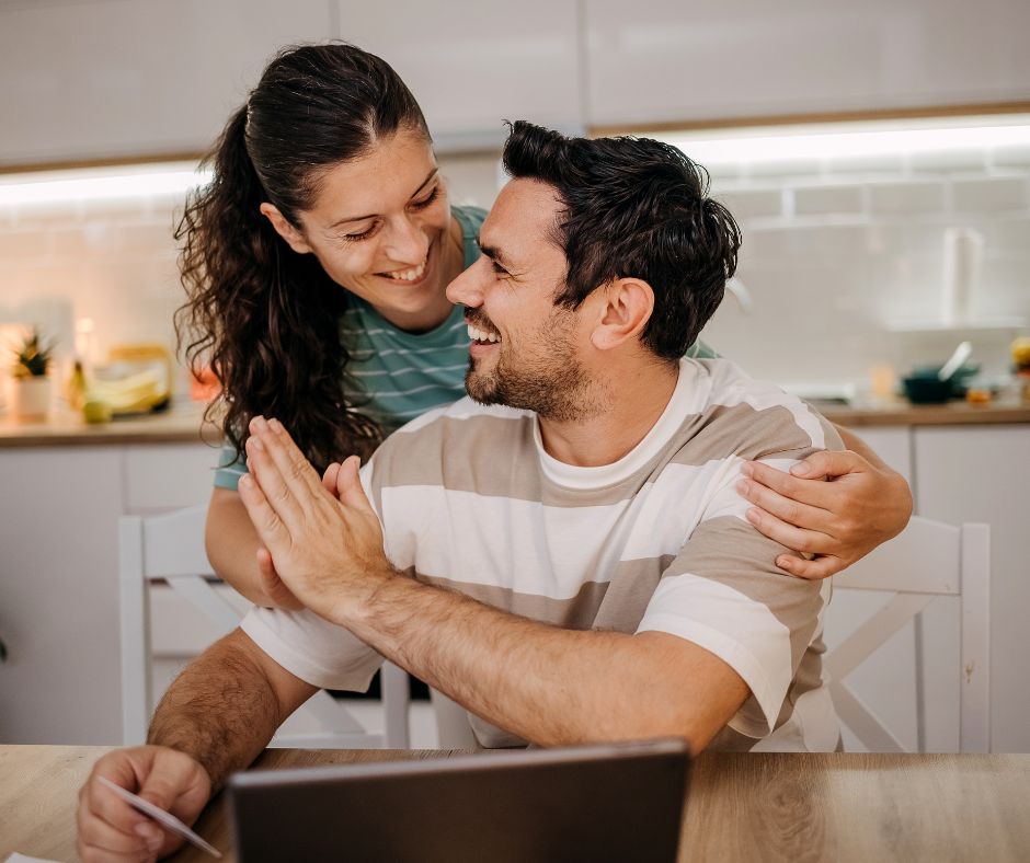 Young couple in white kitchen looking at their computer and high fiving.